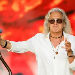 Grzegorz Markowski w białej koszuli i okularach przeciwsłonecznych śpiewa na tle czerwonej sceny.