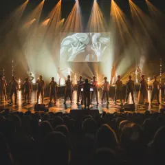Chór męski śpiewający na scenie, oświetlony reflektorami, z ekranem w tle.