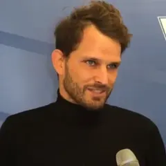 Maciej Synówka, z brodą i w czarnym golfie, udziela wywiadu na tle niebieskiej ściany z logo PZT.