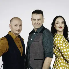 Trzy osoby: łysy mężczyzna w kamizelce, mężczyzna w zielonej koszuli i kamizelce, kobieta w żółtej sukience w grochy.