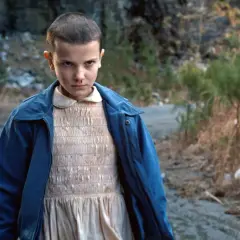 Nastka z Stranger Things, z krótkimi włosami i w niebieskiej kurtce, stoi na brudnej drodze.