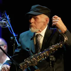 Jan Ptaszyn Wróblewski, legendarny saksofonista, w czapce i garniturze, trzymający saksofon.