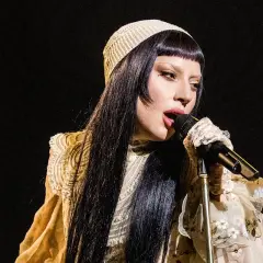 Lady Gaga w beżowym nakryciu głowy i ozdobnej sukni śpiewa do mikrofonu.