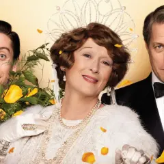 Meryl Streep w koronie i perłach, obok Hugh Granta i Simona Helberga, w scenie z jednego z jej filmów.