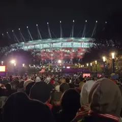 Tłum ludzi zmierza na roztanczony PGE Narodowy. Stadion oświetlony na czerwono, nocna atmosfera.