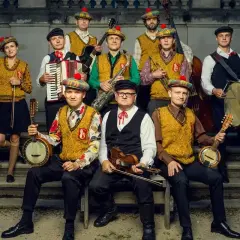 Zespół muzyczny w strojach ludowych z kapeluszami w kratę, grający na instrumentach: banjo, skrzypcach, akordeonie, saksofonie, kontrabasie i bębnie.