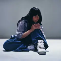 Billie Eilish siedzi na podłodze w luźnych dżinsach i koszuli, patrząc w obiektyw.