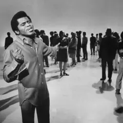 James Brown śpiewa na scenie, otoczony przez publiczność.
