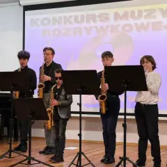 Młodzi muzycy grają na instrumentach: skrzypcach, saksofonach i gitarach, na tle ekranu z napisem "Konkurs Muzyki Rozrywkowej".