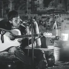 Młody mężczyzna gra na gitarze akustycznej, siedząc na krześle w surowym wnętrzu.