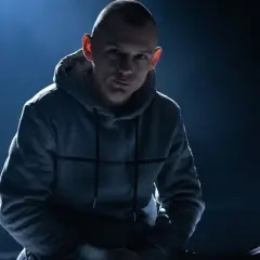 Mężczyzna w szarej bluzie z kapturem, oświetlony od góry, na ciemnym tle.
