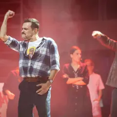 Aktorzy na scenie musicalu, jeden z podniesioną pięścią, w tle inne postacie i napisy.