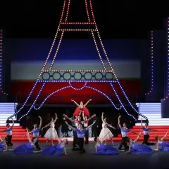 Tancerze baletowi na scenie z miniaturą Wieży Eiffla. W tle czerwono-niebieska scenografia, przypominająca flagę Francji. W powietrzu unosi się aria.