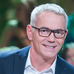 Uśmiechnięty mężczyzna w okularach i granatowej marynarce, z mikrofonem przy uchu.