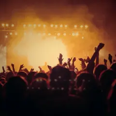 Tłum ludzi z rękami w górze na koncercie, oświetlona scena w tle.