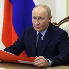 Władimir Putin w garniturze, z rosyjską flagą w tle, trzyma teczkę.
