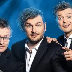 Trzech mężczyzn w garniturach na niebieskim tle, jeden w okularach, jeden z rozczochranymi włosami, jeden wskazujący palcem.