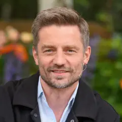 Marcin Rogacewicz, wiek nieznany, uśmiecha się do kamery w otoczeniu zieleni i kolorowych kwiatów.