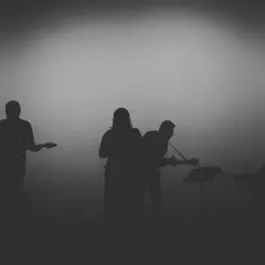 Sylwetki muzyków grających na gitarach i klawiszach na tle mglistego światła.