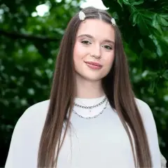 Uśmiechnięta Nicole Pniewska w białej bluzce, z długimi brązowymi włosami i delikatnymi spinkami, na tle zielonych liści.