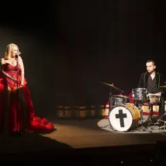 Kasia Gołomska w czerwonej sukni śpiewa przy mikrofonie, a za nią perkusista gra na bębnach.