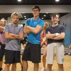 Trzech mężczyzn w strojach sportowych stoi z rękami skrzyżowanymi na piersi, w tle grupa kobiet ćwiczy.