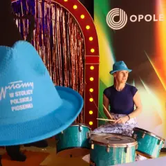 Kobieta w niebieskim kapeluszu gra na perkusji przed sceną z napisem "Opole".