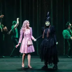 Elphaba w czarnej sukni i Glinda w różowej sukience trzymają się za ręce na scenie.