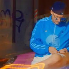 Mężczyzna w niebieskiej kurtce i czapce siedzi na tle nocnego miasta z neonowymi światłami.