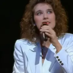 Celine Dion w białej marynarce śpiewa do mikrofonu na ciemnej scenie.