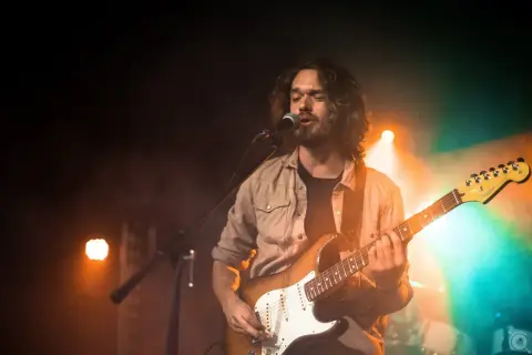 Mężczyzna z długimi włosami gra na gitarze elektrycznej i śpiewa przy mikrofonie na scenie oświetlonej pomarańczowym i zielonym światłem.
