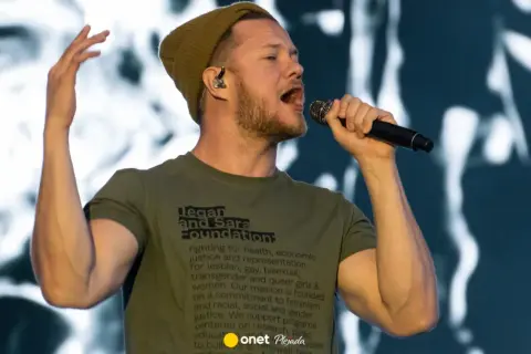 Wokalista Imagine Dragons śpiewa do mikrofonu, ubrany w zieloną koszulkę i czapkę.