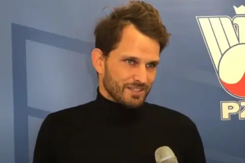 Maciej Synówka, z brodą i w czarnym golfie, udziela wywiadu na tle niebieskiej ściany z logo PZT.
