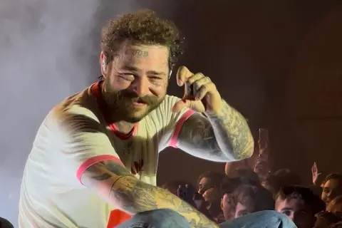 Post Malone na koncercie, uśmiecha się i pokazuje gest ręką.