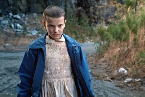 Nastka z Stranger Things, z krótkimi włosami i w niebieskiej kurtce, stoi na brudnej drodze.