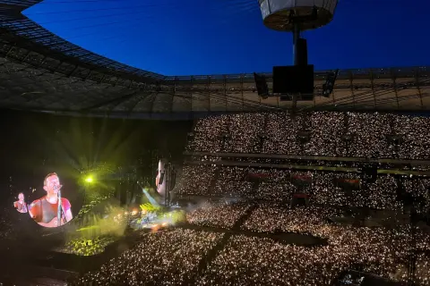 Koncert na stadionie, tłum z zapalonymi telefonami, na ekranie wokalista Coldplay.