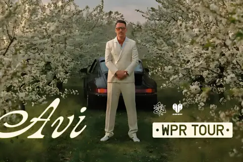 Mężczyzna w garniturze stoi przed czarnym samochodem wśród kwitnących drzew. Napis "Avi" i "WPR TOUR".