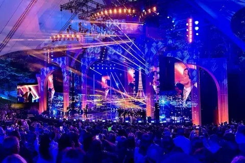 Koncertowa scena z tłumem widzów, oświetlona niebieskimi i różowymi światłami, z ekranami wyświetlającymi obrazy i wieżę Eiffla.