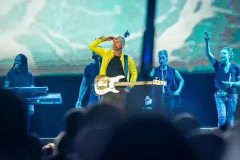 Sting w żółtej kurtce gra na gitarze basowej na scenie z zespołem.