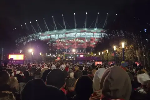 Tłum ludzi zmierza na roztanczony PGE Narodowy. Stadion oświetlony na czerwono, nocna atmosfera.