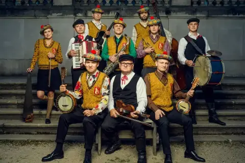 Zespół muzyczny w strojach ludowych z kapeluszami w kratę, grający na instrumentach: banjo, skrzypcach, akordeonie, saksofonie, kontrabasie i bębnie.