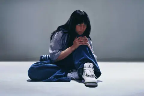 Billie Eilish siedzi na podłodze w luźnych dżinsach i koszuli, patrząc w obiektyw.
