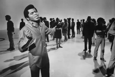 James Brown śpiewa na scenie, otoczony przez publiczność.