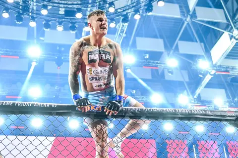 Alan Kwieciński, wytatuowany zawodnik MMA, w rękawicach, oparty o siatkę klatki, w świetle reflektorów.