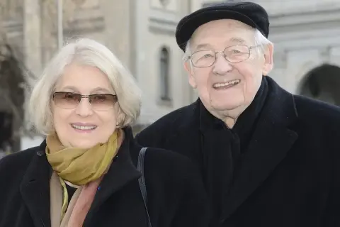 Uśmiechnięta para: Andrzej Wajda i jego żona, w eleganckich płaszczach i szalikach, pozują do zdjęcia na tle architektury.