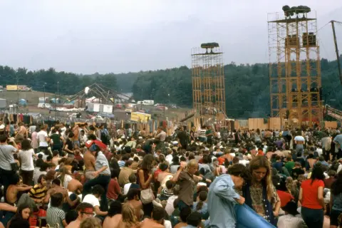 Tłum ludzi na festiwalu, widoczne wieże z głośnikami i scena w tle.