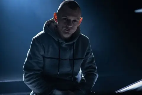 Mężczyzna w szarej bluzie z kapturem, oświetlony od góry, na ciemnym tle.