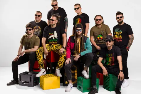 Dziewięciu mężczyzn w okularach przeciwsłonecznych, ubranych w koszulki z motywami reggae, siedzi na skrzynkach po napojach.