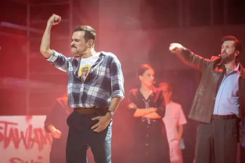Aktorzy na scenie musicalu, jeden z podniesioną pięścią, w tle inne postacie i napisy.