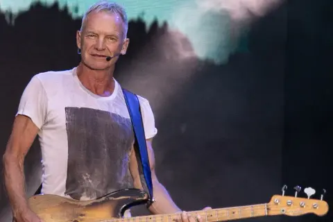 Sting gra na gitarze basowej w białej koszulce na tle sceny.
